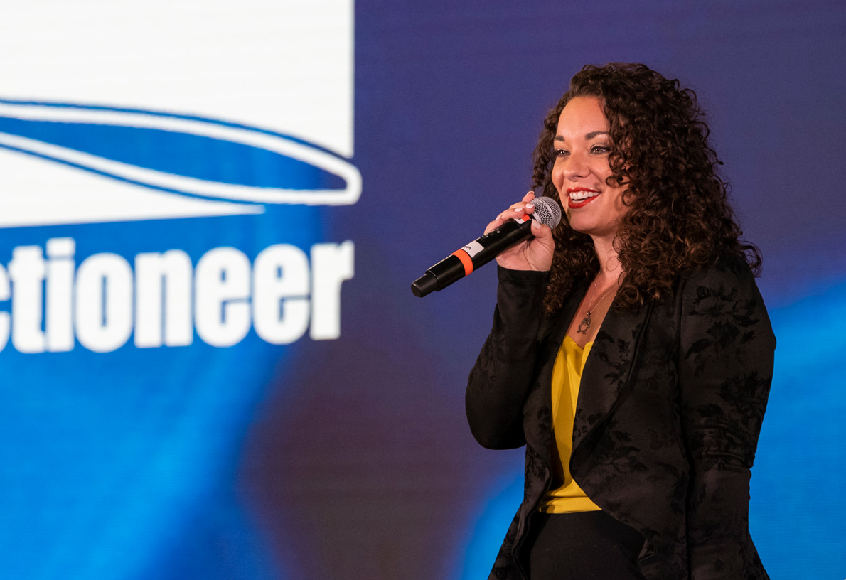 Micki Pickering holding a microphone in front of an auctioneer event banner